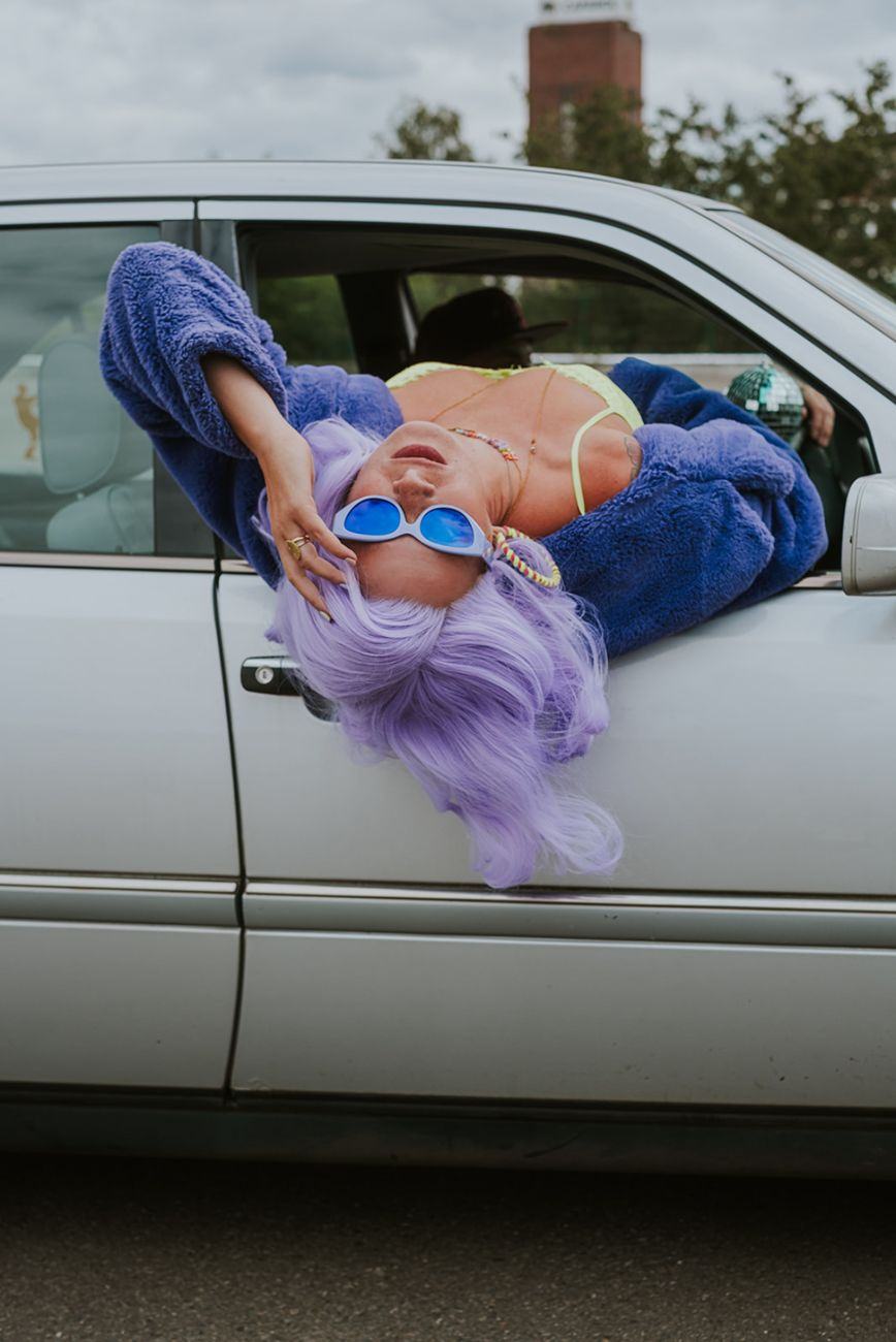 Marie beim Shooting mit lila Perücke, Teddyjacke und blauer Sonnenbrille aus dem Autofenster gelehnt